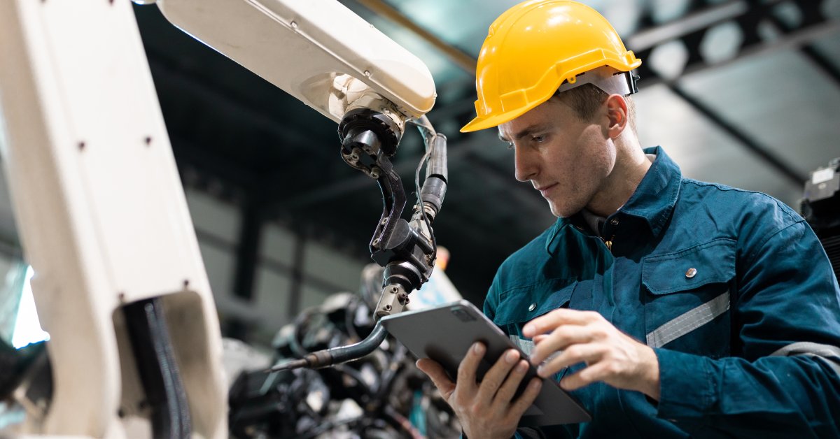 A worker wearing a yellow helmet and a blue uniform holding a tablet and standing in front of a robotic arm in a warehouse.