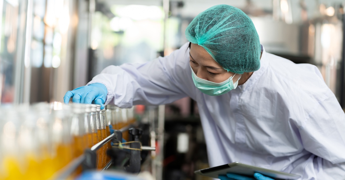 Worker wearing a white robe and safety gloves, touching a bottle filled with juice on a conveyor belt next to other bottles.