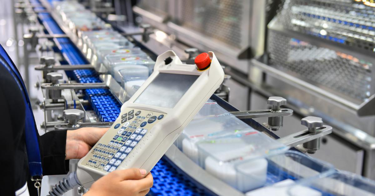 A person holding a large remote control with multiple buttons and a screen over an automated conveyor belt.