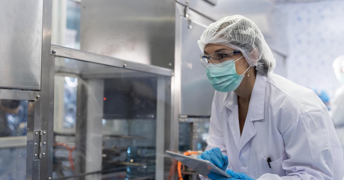 A woman wearing a white coat, safety goggles, face mask, hairnet, and gloves holds a tablet and looks at a machine.