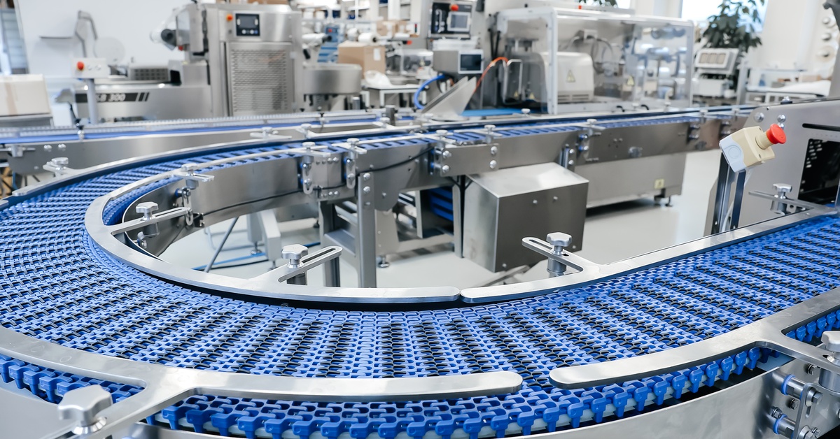 An empty conveyor belt with a blue plastic base formed with tiny locks in a U-shape is inside a large warehouse.