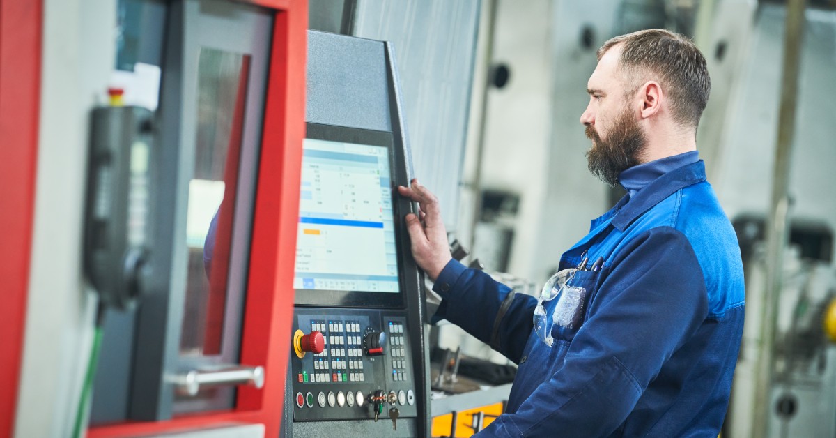 A worker wearing a blue uniform standing in front of a large machine, touching the digital screen with multiple buttons.