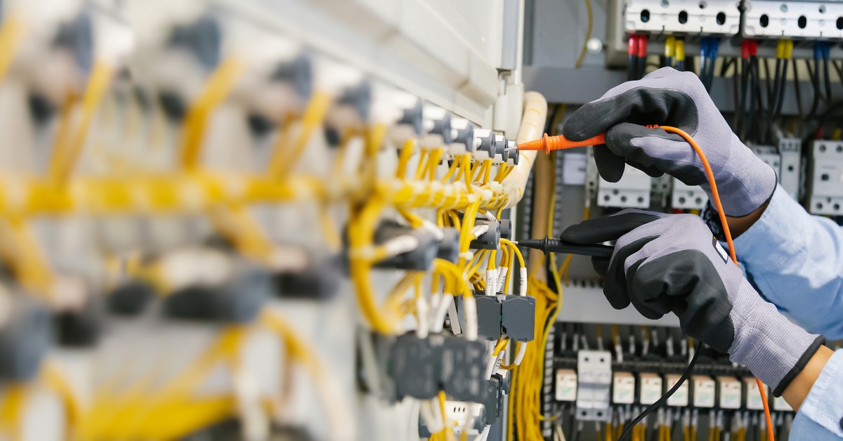Close-up of two hands wearing protective gloves, holding two electrical fusers on a large electrical panel.