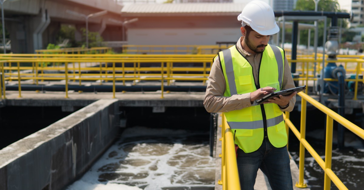 A worker holding a tablet while standing on a bridge on top of a large concrete container with dark water.