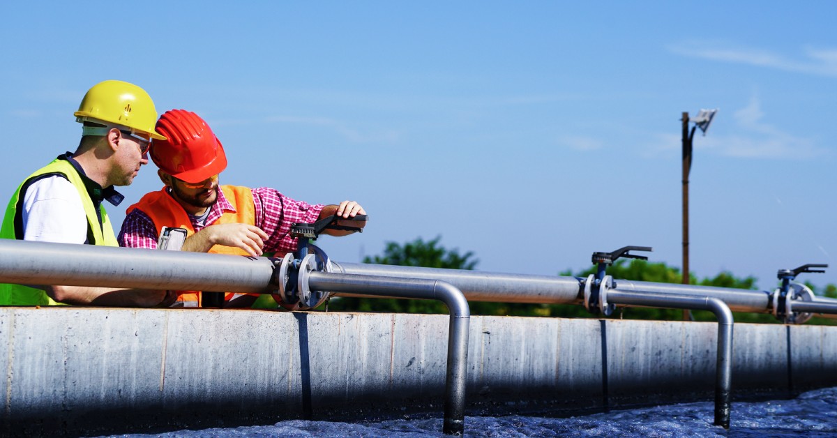 Two workers wearing safety helmets holding a pair of clamps on the connection of multiple metal tubes outdoors.