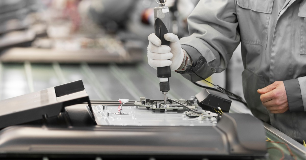 A close-up of the hand of a man using a screwdriver on a product placed in an assembly line inside a large warehouse.