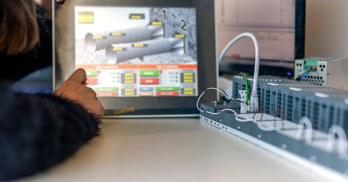 A close-up of the hand of a woman sitting behind a desk, touching a digital screen next to a long electrical panel.