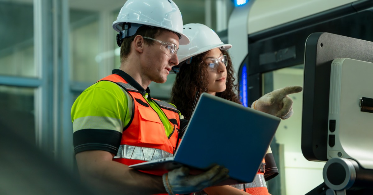 A male and a female worker wearing white helmets and safety vests, looking at a screen while the man holds a laptop.