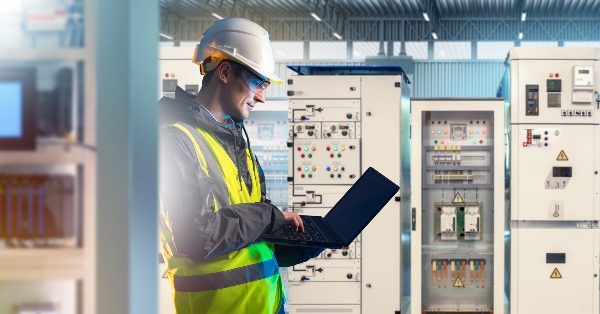 A worker wearing a safety vest and a helmet, holding a laptop inside a room with a large electrical panel in the back.