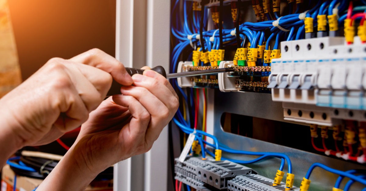 A close-up of two hands holding a screwdriver and touching a wire connection inside a panel with connectors and cables.