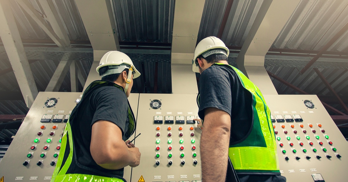 Two workers wearing safety vests and white helmets, standing in front of a large electrical panel with multiple buttons.