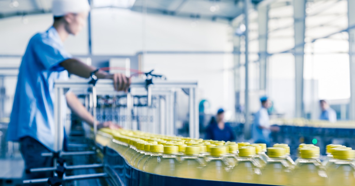 A man wearing a white hat, pointing at a conveyor belt carrying multiple bottles filled with yellow liquid.