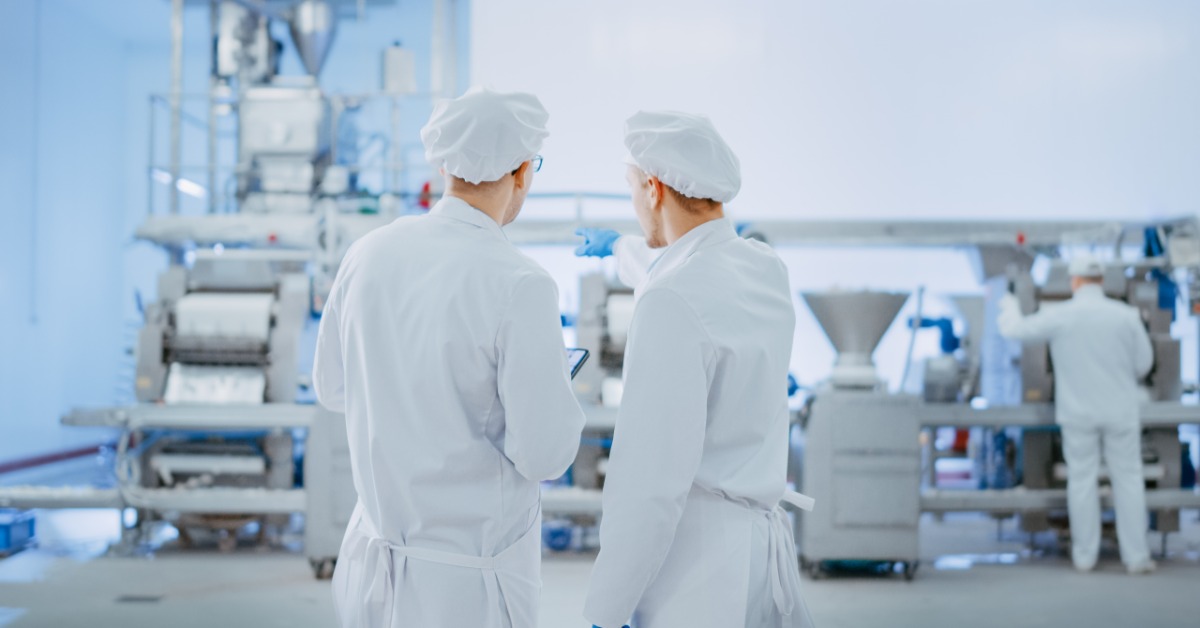 Two workers wearing white hats and uniforms, standing next to each other, looking at a large machine inside a warehouse.