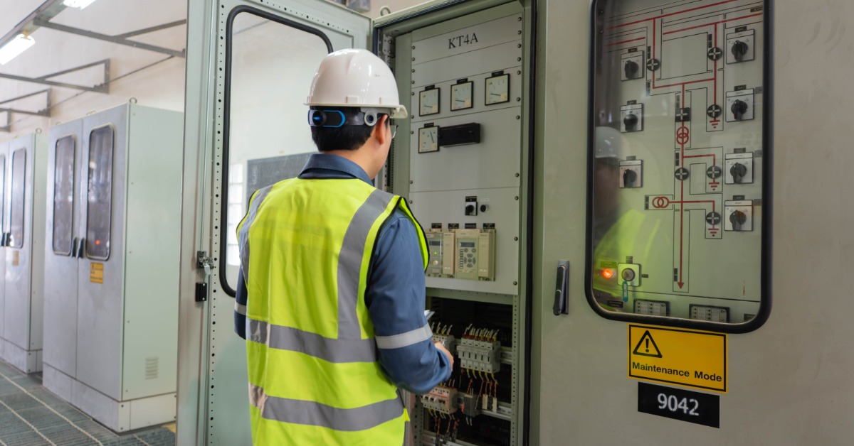 A man wearing a safety vest and a white helmet looking at an electrical panel with multiple connections and buttons.