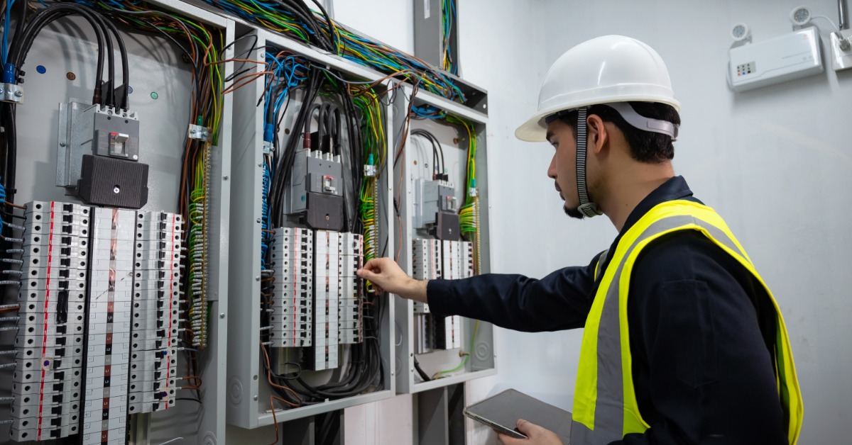 A worker wearing a white helmet, holding a tablet while touching a switch on a large control panel attached to the wall.